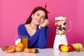 Positive woman with cheerful facial expression, mixes fruit in blender for making smoothie, keeps hands together near face,