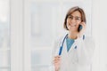 Positive woman doctor consults patient via cellphone, looks gladfully at camera, holds pen, wears white coat, poses in medical Royalty Free Stock Photo