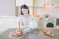 Positive woman sits at table in kitchen. She smiles and poures water into white cup. There are plates with cookies nad Royalty Free Stock Photo
