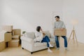 Positive woman and man pose in empty spacious room during relocation day, husband carries cardboard boxes with belongings, wife Royalty Free Stock Photo