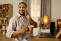 Positive young African American man with coffee talking on cellphone, using laptop computer at home office, copy space Royalty Free Stock Photo