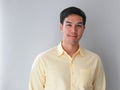 Positive young Asian man in light yellow shirt smiling and looking at camera on grey background, authentic half body portrait.