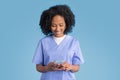 Positive young black lady doctor nurse in blue uniform typing on smartphone, chatting with client Royalty Free Stock Photo