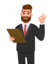 Positive young businessman wearing a suit holding clipboard and showing or making okay,OK gesture or sing with hand fingers. Royalty Free Stock Photo