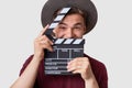 Positive young cameraman holds clapboard near face, has joyful expression, wears hat, prepares for making cutaway, involved in fil