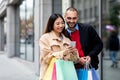 Positive young diverse couple with bright paper bags and cellphone ordering goods online near shopping mall Royalty Free Stock Photo