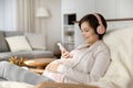 Positive young expectant mom in pink wireless headphones chatting online