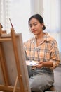 Positive young female artist holding palette and painting picture on canvas with oil paints in her home studio Royalty Free Stock Photo