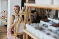 Positive young female model with pink hair sits at wooden easel with canvas