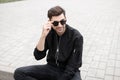 Positive young hipster man with smile in trendy sunglasses in a stylish black jacket in jeans is sitting on the sidewalk Royalty Free Stock Photo