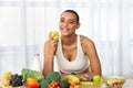 Positive young latin woman in sportswear eat green apple in light kitchen