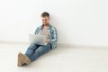 Positive young man in casual clothes surfing the Internet in search of new housing sitting on the floor in an empty room