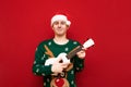 Positive young man in santa hat and green christmas sweater playing on ukulele and looking at camera with smile on face.Cheerful Royalty Free Stock Photo