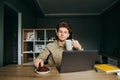 Positive young man in a shirt and wireless headphones sitting at work at home and working on a laptop, taking a break, eating Royalty Free Stock Photo