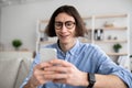 Positive young man sitting on couch with smartphone, chatting with ladies, using dating mobile application on cellphone Royalty Free Stock Photo