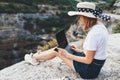 Positive young millennial girl with hat sitting outdoors in mountains working from distance using laptop, beautiful tourist girl Royalty Free Stock Photo