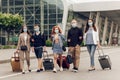 Positive young people in protective masks with luggage outdoor near the airport. Friends returning from a trip.