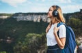 Positive young traveler girl with headphones and backpack stands on top of mountain, active tourist woman hiking enjoys beautiful