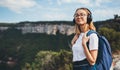 Positive young traveler girl with headphones and backpack stands on top of mountain, active tourist woman hiking enjoys beautiful