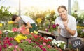 Positive young woman choosing potted Petunia flowers for house while shopping in greenhouse