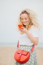 Positive young woman uses modern smart phone, has cheerful expression, makes shopping online, carries bright small bag, has outdoo Royalty Free Stock Photo