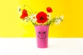 postcard a bouquet of daisies and poppies in a pink vase with a painted muzzle on the table on a yellow background. Royalty Free Stock Photo