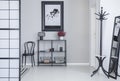 Poster above shelves with flowers and lamp in white simple hall interior with rack and black chair