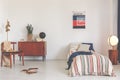 Poster above striped bed in white vintage bedroom interior with chair next to wooden cabinet. Real photo Royalty Free Stock Photo