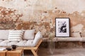 Poster and flowers next to grey wooden couch with pillows in flat interior with brick wall