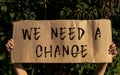 A poster with the words WE NEED A CHANGE in the hands of a young girl on a background of green foliage