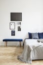 Posters above blue bench next to bed with grey blanket in white simple bedroom interior. Real photo