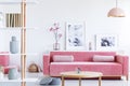 Posters and flowers above pink sofa in pastel living room interior with wooden table. Real photo