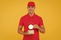 Postman delivery worker. Handsome man red cap hold alarm clock yellow background. Delivering purchase. Open hours. Time