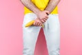 Potency and men`s health. A man in white jeans, legs apart, holds a banana near the genitals. Pink background. Close up of hands