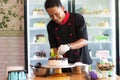 Potrait of asian male pastry chef in a black uniform decorating round vanila cake with melted chocolate and red cherry