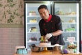 Potrait of asian male pastry chef in a black uniform decorating round vanila cake with melted chocolate and red cherry