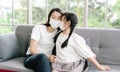 Potrait of happy young Asian mother and little daughter with protective medical face masks on sitting sofa and hugging during