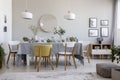 Pouf, plants and round mirror in bright dining room interior with chairs at table under lamps