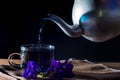 Pour Butterfly pea tea by metal pot to cup with violet flower on brown table cloth on black background. Healthy beverrage for Royalty Free Stock Photo