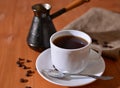 poured black coffee into a white cup near the turk and coffee grains on burlap and wooden table