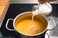 Pouring coconut milk or cream in a vegetable soup from red kuri squash or Hokkaido pumpkin in a steel pot on a black stove, Royalty Free Stock Photo