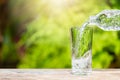 Pouring drinking water from bottle into glass on wooden tabletop on blurred fresh green nature background