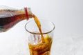 Pouring fresh cola from a bottle into a drinking glass with ice cubes and lemon slice, refreshing drink against a light gray Royalty Free Stock Photo