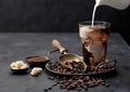 Pouring fresh milk into glass of iced black coffee on tray with beans and ground coffee with cane sugar and vintage shop on black Royalty Free Stock Photo