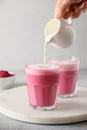 Pouring frothed milk in to beetroot latte in glass on gray background.