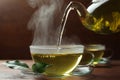 Pouring green tea into glass cup with saucer and leaves on wooden table, closeup Royalty Free Stock Photo