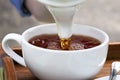 Pouring high quality hot tea from an elegant white teapot to a traditional white cup on an organic wooden tray. Extreme closeup