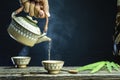 pouring hot tea from a teapot into cup steam is over the cupping, spoon, bamboo leaves, wooden vase on the old wood table a black Royalty Free Stock Photo