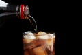 Pouring refreshing cola from bottle into glass with ice cubes on black background