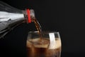 Pouring refreshing cola from bottle into glass with ice cubes on black background